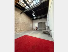 Leere Industriehalle mit hoher Decke, sichtbaren Stahlträgern und Oberlichtern; links eine freiliegende Ziegelwand und rechts weiße Wände. Am Ende befinden sich ein großes Rolltor, ein kleines Fenster und eine Metallplattform mit Treppe; im Vordergrund liegt ein roter Teppich auf dem Betonboden.