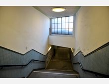 Eine Treppenunterführung mit hellen Wänden und blauen Fliesen an den Seitenwänden. Oben befindet sich ein großes Fenster mit Glasbausteinen, das Tageslicht einfällt; der Boden und die Wände zeigen Gebrauchsspuren.