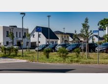 Moderne Wohnsiedlung mit Einfamilienhäusern in hellen Farben und dunklen Dächern, davor ein Parkplatz mit mehreren Autos. Im Vordergrund befindet sich eine Grünfläche mit jungen Bäumen, sowie Straßenschilder für die Straßen "Balzacring" und "Rousseauallee".