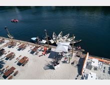 Ein Strandbereich mit Tischen und Stühlen auf hellem Sand, angrenzend an einen ruhigen Fluss, an dessen Ufer mehrere Ruderboote und Kanus festgemacht sind. Ein Sonnenschirm steht in der Mitte, und Paddel sind in einem Ständer organisiert.