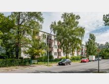 Mehrstöckiges Wohngebäude mit hellen Fassaden und Balkonen, umgeben von hohen Bäumen und Grünflächen. Im Vordergrund verläuft eine Straße mit parkenden Autos und einem Straßenschild "Fercher Straße".