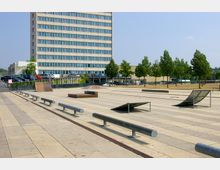Ein moderner, gepflasterter Skatepark mit Metall-Rails und Rampen, umgeben von einer Reihe junger Bäume. Im Hintergrund steht ein mehrstöckiges Bürogebäude mit vielen Fenstern, daneben ist ein Parkplatz mit Autos zu sehen.