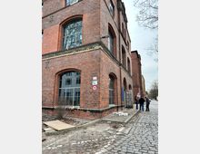 Ehemaliges Industriegebäude aus roten Backsteinen mit großen, bogenförmigen Fenstern. Der Bereich um das Gebäude zeigt eine Kopfsteinpflasterstraße und unbefestigte Flächen; im Vordergrund stehen drei Personen.