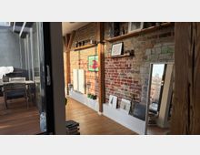 Wohnraum mit einer freigelegten Ziegelwand, Holzbalken und dekorativen Regalen, auf denen Bilder und Dekorationsgegenstände stehen. Eine Glastür führt zu einer Holzterrasse mit Tisch und Stühlen.