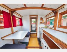 Innenraum eines schmalen Wohnwagens mit Holzelementen, einer Sitzecke mit weißem Klapptisch und rotem Vorhang links, einer Küchenzeile mit Spüle und Arbeitsfläche rechts sowie einem schmalen Gang in der Mitte, der zu weiteren Bereichen führt.