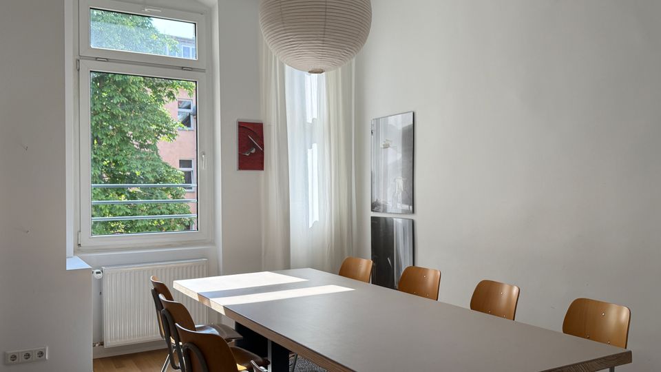 Heller Besprechungs- oder Essraum mit langem rechteckigem Tisch und mehreren Holzstühlen, darüber eine große runde Papier-Hängelampe. Links ein großes Fenster mit Blick auf Bäume und ein gegenüberliegendes Gebäude, an der Wand hängen zwei gerahmte Bilder.