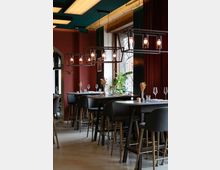 Ein modernes Restaurant mit hohen Tischen und Barhockern, dunkelroten Wänden und großen Fenstern, die Tageslicht hereinlassen. Industrielle Pendelleuchten und vereinzelte Pflanzen betonen die stilvolle Einrichtung.