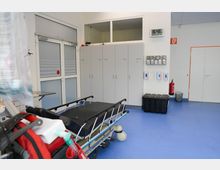 Ein steriler Krankenhausraum mit blauen Böden, einem fahrbaren Krankenbett im Vordergrund und Schränken an der Wand. An der rechten Wand befinden sich ein Feuerlöscher, Spender für Hygienehandschuhe und ein Wanduhrenanzeige mit Uhrzeit und Datum.