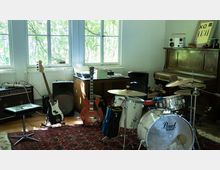 Musikraum mit Tageslicht, eingerichtet mit verschiedenen Instrumenten: E-Gitarre, Bass, Schlagzeug und einem Klavier. Im Hintergrund befinden sich große Fenster, Lautsprecher, ein Teppich und Möbel mit weiterem Zubehör.