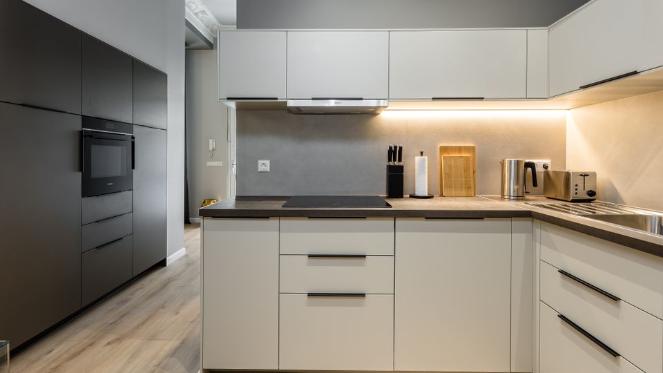 Moderne, L-förmige Einbauküche mit hellen Fronten, dunkler Arbeitsplatte und grauer Rückwand, beleuchtet durch eine durchgehende Unterbauleuchte. Links stehen hohe schwarze Schrankelemente mit integriertem Backofen; rechts sind Spüle, Toaster und Wasserkocher auf der Arbeitsfläche.