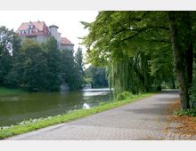 Uferweg mit Bäumen und herabhängenden Weiden entlang eines Flusses, im Hintergrund ein mehrstöckiges Gebäude mit rotem Ziegeldach, umgeben von dichtem Grün. Entlang des Flusses verläuft ein gepflasterter Weg mit Laub auf dem Boden.