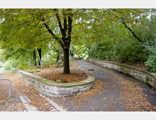 Weg in einem Park mit gepflasterten Gehwegen, flankiert von Steinmauern. Die Umgebung ist von dichtem Laub und großen Bäumen geprägt, und der Boden ist mit Herbstlaub bedeckt.