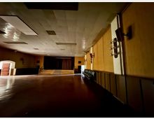 Ein leerer Saal mit einem glänzenden Fußboden, einer kleinen Bühne im Hintergrund und holzverkleideten Wänden. Rechts entlang der Wand befindet sich eine Reihe von Wandlampen und Stühlen, links ist ein geöffneter Türbogen sichtbar.