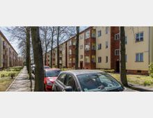 Straßenszene mit einer Reihe von dreistöckigen Wohngebäuden im Stil der Nachkriegszeit, mit gelben Fassaden und roten Klinkerdetails. Im Vordergrund stehen parkende Autos, und ein Gehweg mit Bäumen verläuft entlang der Gebäude.