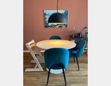 Runder Esstisch mit weißer Tischplatte, umgeben von drei blauen Polsterstühlen und einem weißen Kinderstuhl, auf einem Holzboden. Darüber hängt eine schwarze, halbkugelförmige Lampe, im Hintergrund eine rotbraune Wand mit einem Landschaftsbild und einem Regal mit Dekoration.