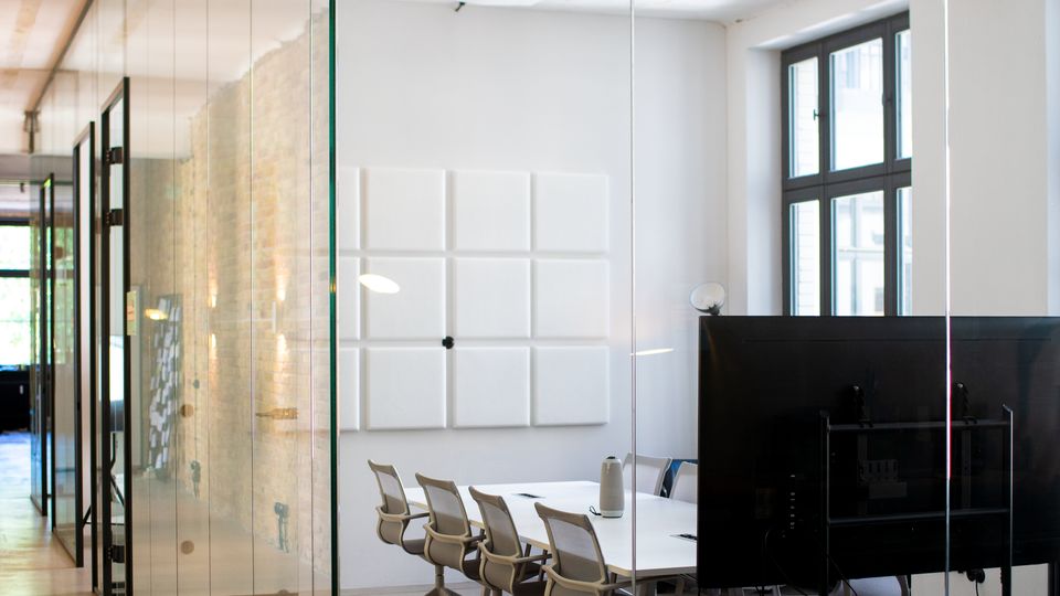 Moderner Konferenzraum in einem Büro, durch eine raumhohe Glaswand vom Flur getrennt. Im Raum stehen ein langer Tisch mit mehreren Stühlen, große Fenster, eine Decke mit quadratischen Akustikpaneelen sowie ein grünes Notausgangsschild über der Tür.