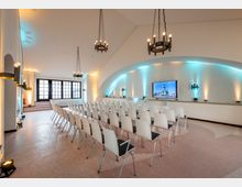 Heller Konferenzraum mit weißen Wänden, modernen Stuhlreihen und einem großen Bildschirm an der beleuchteten Stirnwand. Über den Sitzplätzen hängen schwarze Kronleuchter, und ein großes Fenster mit dunklen Rahmen befindet sich im Hintergrund.