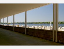 Überdachter Gehweg mit Blick auf einen Sandstrand mit nummerierten Strandkörben, einem See im Hintergrund und einer bewaldeten Uferlandschaft.