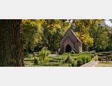 Kleine Friedhofskapelle mit spitzem Ziegeldach und einem Kreuz über dem Eingang, umgeben von gepflegten Grünflächen, Gräbern und Bäumen in einer parkähnlichen Umgebung. Im Vordergrund sind ein breiter Baumstamm und blühende Büsche zu sehen.