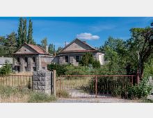 Verlassenes Gebäude mit stark verwitterter Fassade, roten Ziegeldächern und vereinzelten Fenstern, umgeben von dichtem Bewuchs und einer rostigen Metallzaunanlage im Vordergrund. Im Hintergrund sind Bäume und ein klarer blauer Himmel sichtbar.