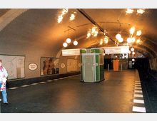 U-Bahn-Station mit gewölbter, gekachelter Decke, beleuchtet durch runde, hängende Lampen. In der Mitte des Bahnsteigs befindet sich ein grüner Kiosk, und an den Wänden hängen Wandbilder und Stationsschilder.