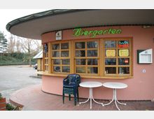 Kleines Biergarten-Gebäude mit runden, überdachten Außenbereich, großen Holzfenstern und einer neon-grünen Schrift mit dem Wort „Biergarten“ an der Fassade. Vor dem Gebäude stehen weiße Tische und gestapelte Plastikstühle auf einem gepflasterten Boden.