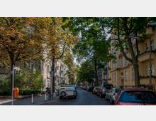 Eine ruhige Wohnstraße mit mehrstöckigen Wohngebäuden auf beiden Seiten, teils in heller und teils in gelblicher Fassade. Die Straße ist von Bäumen gesäumt, deren Blätter teils grün und teils herbstlich gefärbt sind. Geparkte Autos stehen entlang der Straße, und ein Anhänger ist am rechten Straßenrand abgestellt.