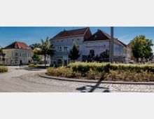 Eine Straßenkreuzung in einer Kleinstadt mit gepflastertem Gehweg und einem kleinen bepflanzten Kreisverkehr. Im Hintergrund stehen Gebäude mit Satteldächern und Schaufenstern, darunter eines mit einer Werbefläche, die den Schriftzug „Willkommen in Wittenberge – Das Tor zur Elbtalaue“ zeigt.