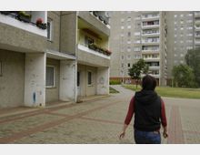 Wohnanlage mit Plattenbauten in einem Wohngebiet. Im Vordergrund sind ein Gehweg, bepflanzte Balkone und eine Person von hinten sichtbar, im Hintergrund sind weitere Wohngebäude und Grünflächen zu sehen.