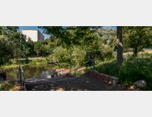 Treppenabgang aus rotem Backstein mit Geländern führt zu einem kleinen Gewässer, umgeben von dichter Vegetation. Im Hintergrund ist ein mehrstöckiges Gebäude hinter Bäumen sichtbar.