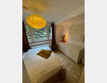 Ein kleines Schlafzimmer mit einem großen Fenster mit Blick auf einen Gartenbereich mit Felsen. Im Raum befinden sich ein Bett mit weißer Bettdecke, eine weiße Kommode, eine dekorative Wandlampe in warmem Gelbton und rote Vorhänge.