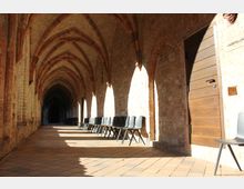 Gewölbegang eines historischen Gebäudes mit gotischen Bögen und hellem Mauerwerk. Entlang der rechten Wand stehen graue Kunststoffstühle unter einem Lichteinfall; eine Holztür mit Beschriftung "Ausstellung" ist ebenfalls sichtbar.