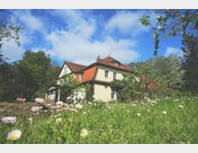 Ein Landhaus mit cremefarbener Fassade und rotem Ziegeldach, umgeben von einer grünen Wiese mit blühenden Gänseblümchen und Bäumen im Hintergrund unter einem teilweise bewölkten blauen Himmel.