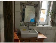 Badezimmer mit weiß gefliesten Wänden, einem Holzwaschtisch mit einem rechteckigen Waschbecken und einem großen, dekorativen Spiegel. Rechts daneben ein Fenster mit gekipptem Flügel und einer Topfpflanze auf der Fensterbank.