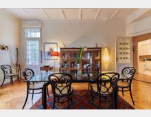 Esszimmer mit Holzboden, Glastisch und sechs Stühlen mit geschwungenen schwarzen Metallrahmen. Im Hintergrund ein hölzerner Vitrinenschrank, eine Stehlampe sowie eine Tür, die in eine angrenzende Küche führt; der Raum wird durch Tageslicht und eine Tischlampe beleuchtet.