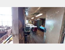 Blick in einen alten, abgenutzten Raum mit der Aufschrift „Meisterstube PS4“ auf der offenen Tür. Der Raum enthält einen Tisch mit Stühlen, Schränke, Aktenordner und Regale, sowie sichtbare Gebrauchsspuren wie abblätternde Farbe an der Wand.