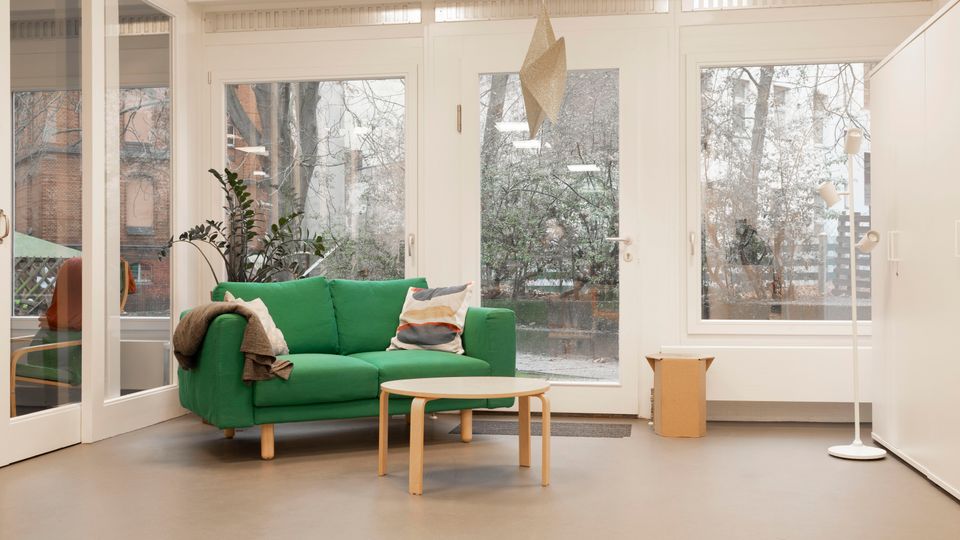Heller Aufenthaltsraum mit großem Fenster- und Türelement zur Außenseite, grauem Boden und weißer Decke. In der Mitte stehen ein grünes Sofa mit Kissen, ein runder Couchtisch sowie eine Stehlampe und eine Hängelampe; draußen sind Bäume und Nachbargebäude zu sehen.