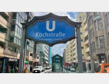 Straßenszene mit einem blauen Schild der U-Bahn-Station "Kochstraße – Checkpoint Charlie" in Berlin, umgeben von mehrstöckigen Wohn- und Geschäftsgebäuden. Im Hintergrund ist der Zugang zur Station mit Glaskonstruktion sichtbar, sowie geparkte und fahrende Autos.