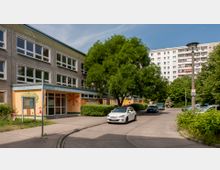 Zweigeschossiges Gebäude einer Kita mit großen Fenstern und einer mit bunten Bildern bemalten Fassade, davor ein gepflasterter Weg mit parkenden Autos. Im Hintergrund sind Grünflächen und ein mehrstöckiges Wohngebäude sichtbar.