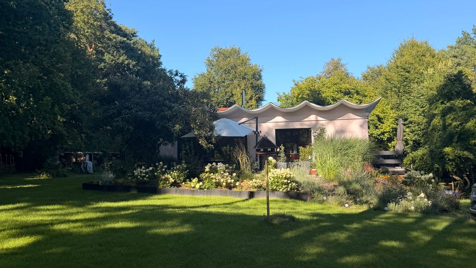 Große Rasenfläche in einem parkähnlichen Garten, umgeben von hohen Bäumen und Beeten mit blühenden Sträuchern. Im Hintergrund steht ein niedriges Gebäude oder Pavillon mit wellenförmigem Dach und einer Terrasse.