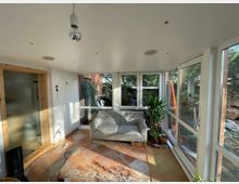 Wintergarten mit großen Fenstern, die viel Tageslicht hereinlassen, ausgestattet mit einem grauen Sessel, mehreren Topfpflanzen und einem braun-beige gefliesten Boden. Auf der linken Seite befindet sich eine Tür mit mattem Glas und Holzrahmen.