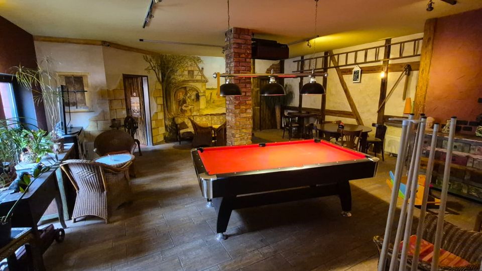 Innenraum eines rustikalen Aufenthalts- oder Gastraums mit rotem Billardtisch unter drei Hängelampen, Backsteinsäule und Holzbalken an den Wänden. Links stehen Pflanzen und Korbsessel an einer Fensterfront, rechts sind Tische, Stühle und ein Billardqueue-Ständer vor einer gemauerten Theke.