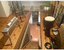 Holzgalerie in einem Innenraum mit hölzernem Fußboden und Treppen. Auf der Galerie stehen ein Trommelinstrument, mehrere Mikrofonständer, eine eingepackte Holzbank, Topfpflanzen und weiteres Zubehör wie Kabel und Kartons.