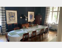 Esszimmer mit einem ovalen Tisch, der mit einer gestreiften Tischdecke gedeckt ist, umgeben von Holzstühlen. Der Raum hat eine dunkelblaue Wand, Holzfußboden, große Fenster mit gestreiften Vorhängen und gerahmte abstrakte Kunstwerke an der Wand.