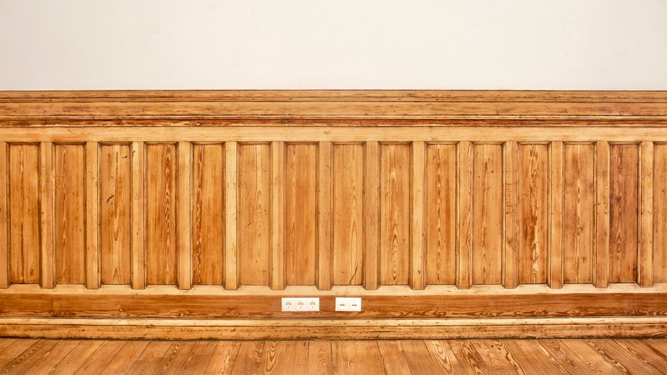 Innenansicht eines Raums mit hoher Holzvertäfelung aus senkrechten Paneelen und breiter Sockelleiste, darüber eine glatte weiße Wand. Vor der Wand liegt ein Holzdielenboden; in der Mitte sind mehrere Steckdosen in die Vertäfelung eingelassen.