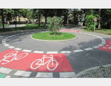 Kreisverkehr für Fahrräder in einem Park, mit einer zentralen Grünfläche und Fahrradwegmarkierungen auf rotem Untergrund. Der Bereich ist von Bäumen und grünem Rasen umgeben, und ein Verkehrsschild weist auf Fußgänger hin.