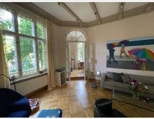 Wohnzimmer mit hohen Decken und dekorativen Holzbalken, großen Fenstern mit Buntglas-Elementen und hellem Vorhang. Im Vordergrund stehen ein graues Sofa und ein Couchtisch mit Blumen, an der Wand hängt ein farbenfrohes Gemälde von Personen mit Regenschirmen. Im Hintergrund führt eine Türöffnung zu einem weiteren Raum mit ähnlicher Einrichtung.