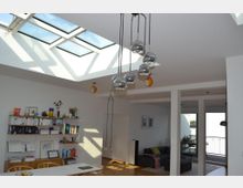 Helles Wohnzimmer mit großer Glasdecke, die viel Tageslicht hereinlässt. Im Raum befinden sich ein Regal mit Büchern und Dekoration, ein Esstisch sowie eine moderne Sofa-Ecke; im Hintergrund ist ein Balkon sichtbar.
