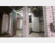 Industrieller Keller mit weißen und teilweise bemalten Ziegelwänden (rot und grün) sowie mehreren Betonpfeilern. In der Mitte führen einfache Holzbretter als Treppe in einen höheren Bereich.