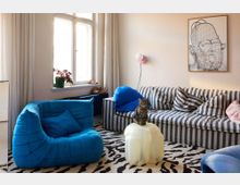 Bright living room with tall windows and light curtains, featuring a striped sofa, a blue lounge chair, and a translucent round coffee table on a black-and-white patterned rug. A wall sconce and framed artwork hang above the sofa, with a radiator and potted plant beneath the window.